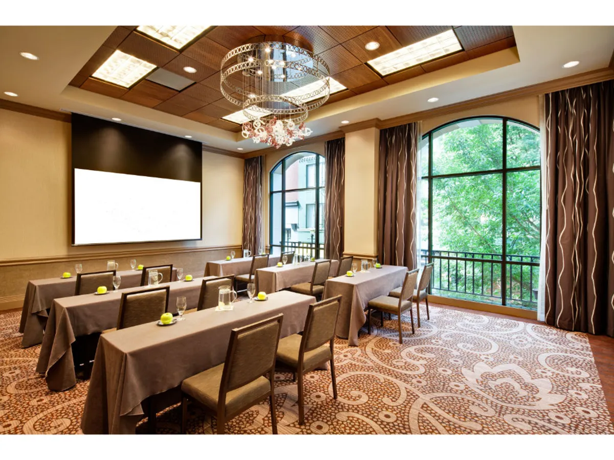 A modern, well-lit meeting space featuring a chandelier, large windows that hint at city views, and tables arranged for business meetings or collaborative workshops. A presentation screen is visible, emphasizing a professional environment geared toward productivity and team engagement.