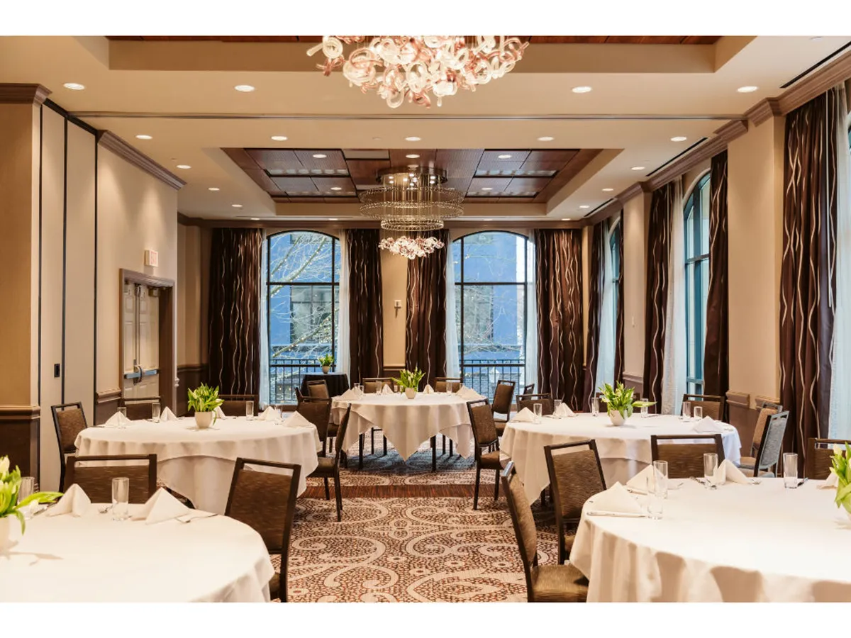 A sophisticated interior features a grand chandelier, white tablecloth tables, decorative plants, and expansive windows. The space is warm and refined, ideal for memorable gatherings, special events, or simply relaxing in style.