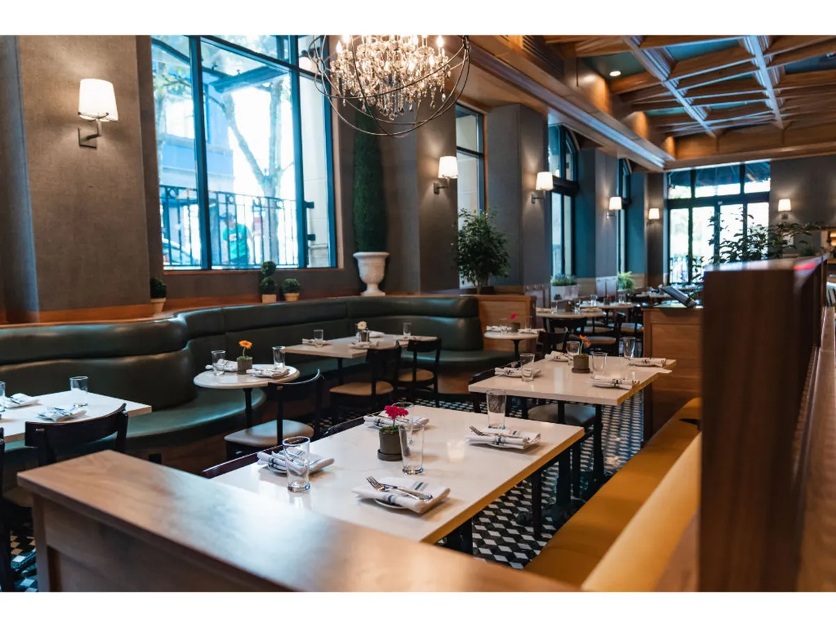 A stylish dining area with elegant green booth seating, large windows letting in natural daylight, and tables adorned with utensils and floral arrangements. The chandelier overhead adds to the inviting yet refined ambiance, perfect for socializing or after-hours relaxation.