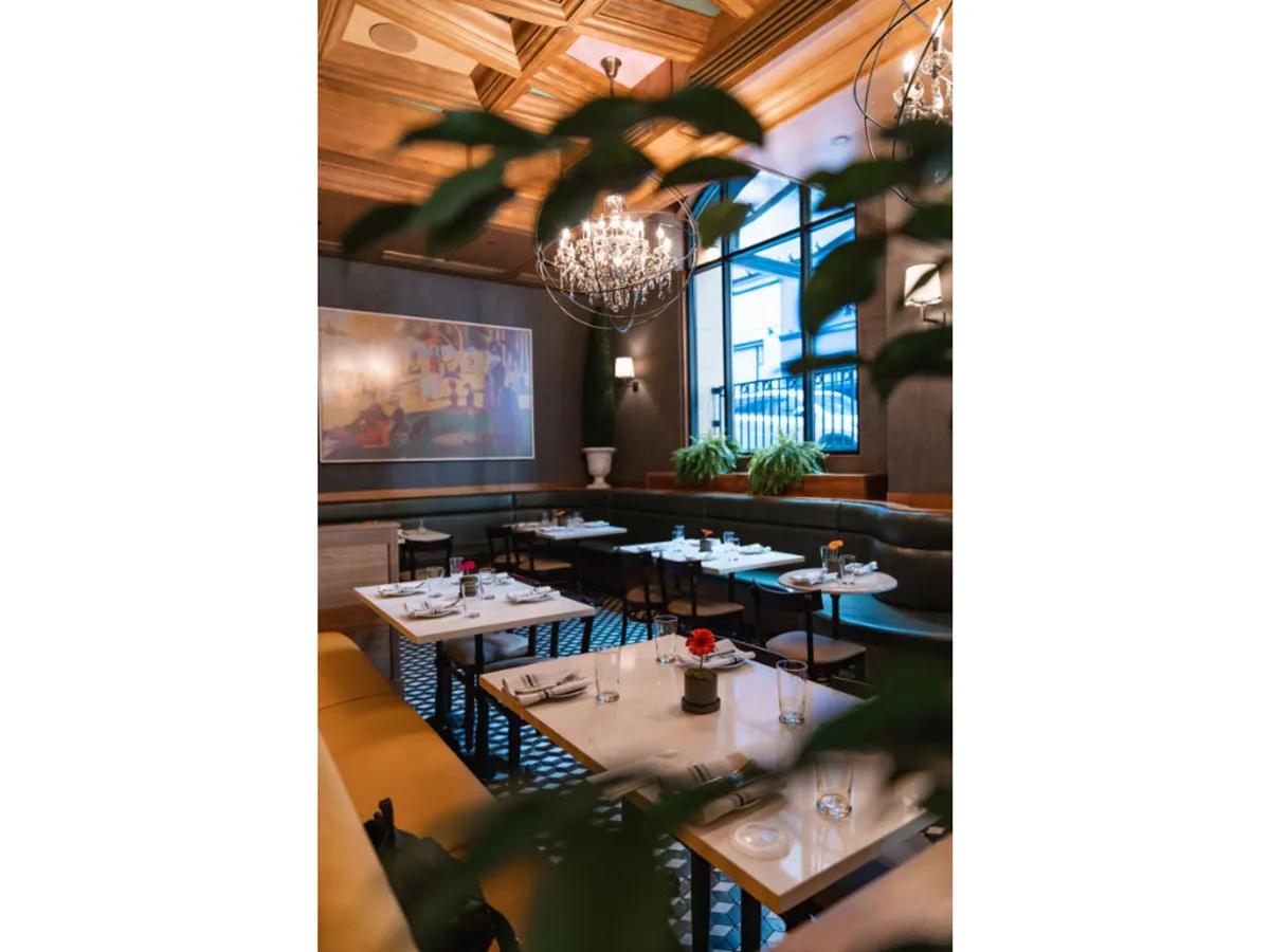 An elegant guestroom or dining area is highlighted by a striking chandelier, modern artwork, and a mix of cozy tables and potted plants near the window. The setting is intimate yet flexible, suitable for everything from solo relaxation to family gatherings.