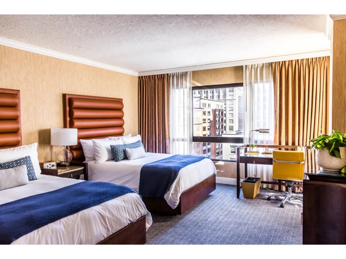 The image shows a bright, inviting hotel room with two stylish beds featuring decorative headboards. A spacious window offers a sweeping view of Portland’s cityscape, while a sleek desk and chair provide a comfortable spot to work or unwind.