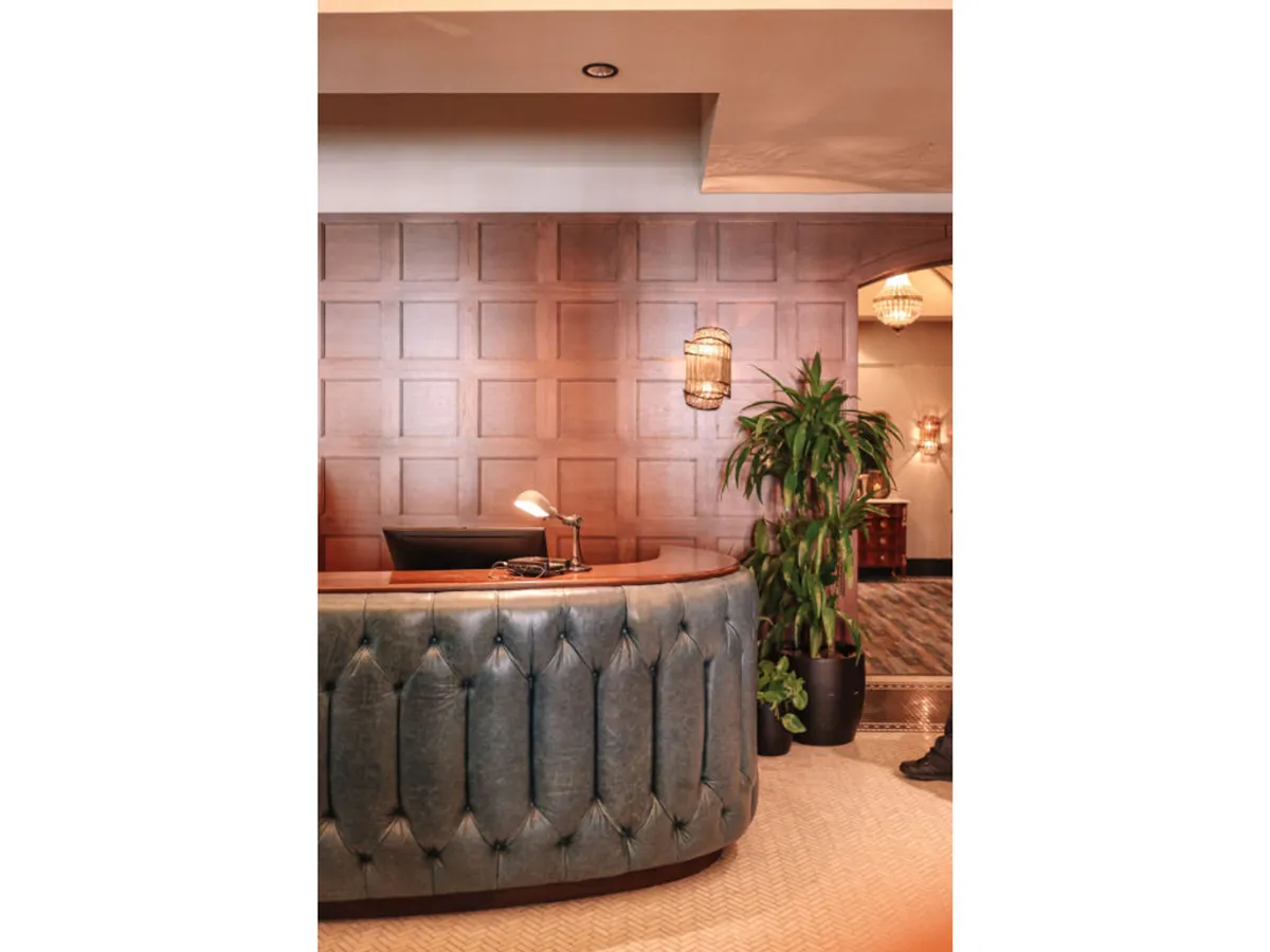 This image shows a modern hotel lobby with a curved reception desk set against a wood panel wall, complemented by hanging light fixtures and a lush indoor plant. The friendly, professional atmosphere is enhanced by guests interacting with staff and relaxing in the inviting space.
