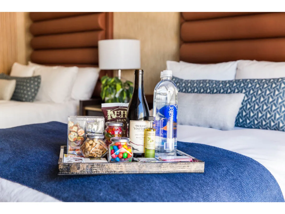 A cozy guestroom scene featuring a tray with snacks and drinks atop a carefully made bed. The bed has inviting linens and decorative pillows, conveying comfort and relaxation. Soft lighting and plush textiles create a welcoming atmosphere, with hints of in-room amenities visible.