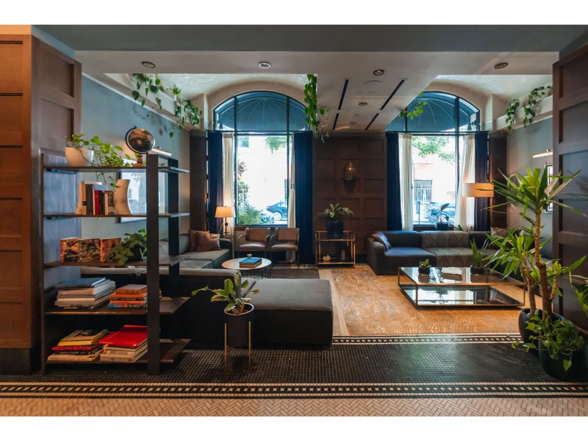 A cozy common area featuring large windows, comfortable seating, a shelf with books and plants, and a coffee table. The space is designed for relaxation, reading, socializing, or enjoying a quiet moment with street views.