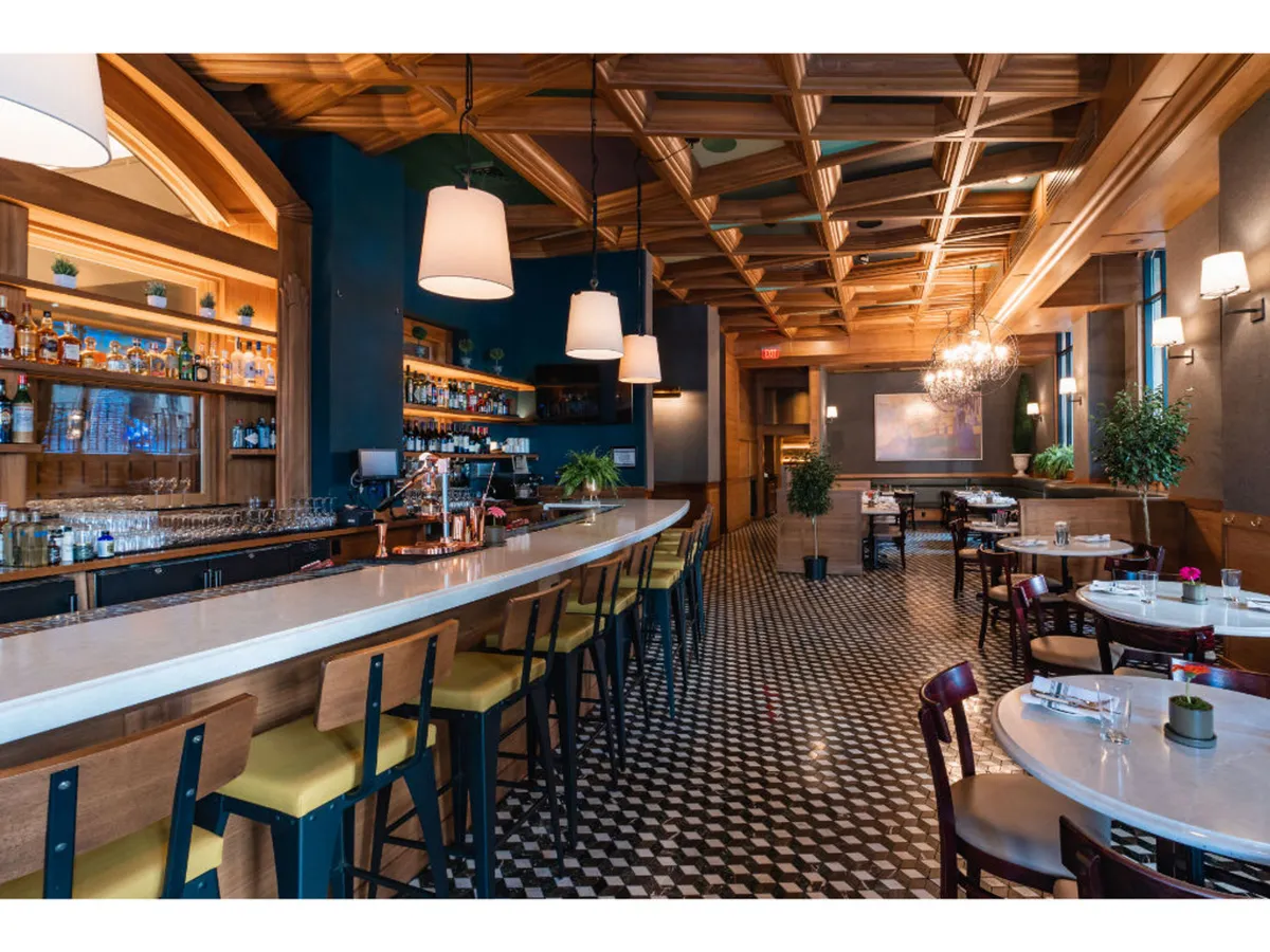 A vibrant interior view of Bar Alder, featuring a stylish bar area, elegant chandeliers hanging from a rich wooden ceiling, and tables thoughtfully set for guests. The space conveys an inviting, lively ambiance ideal for enjoying cocktails or a meal before heading out to explore the city.